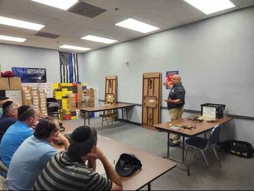 A man gives a presentation to a seated audience in a room. An American flag is visible on the wall.