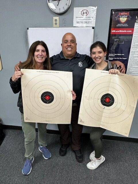 Three people smiling, holding target practice sheets. Two women and a man in a shooting range setting.