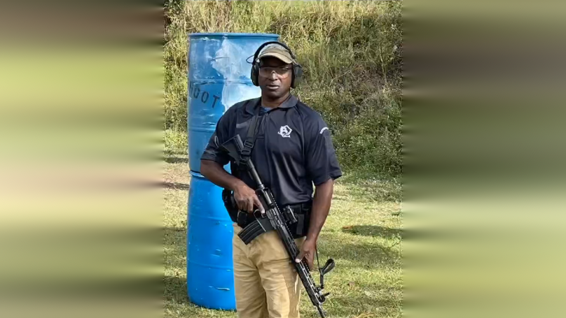 A Black man wearing a dark shirt and tan pants stands with an AR-15 rifle at a shooting range, a blue barrel behind him.
