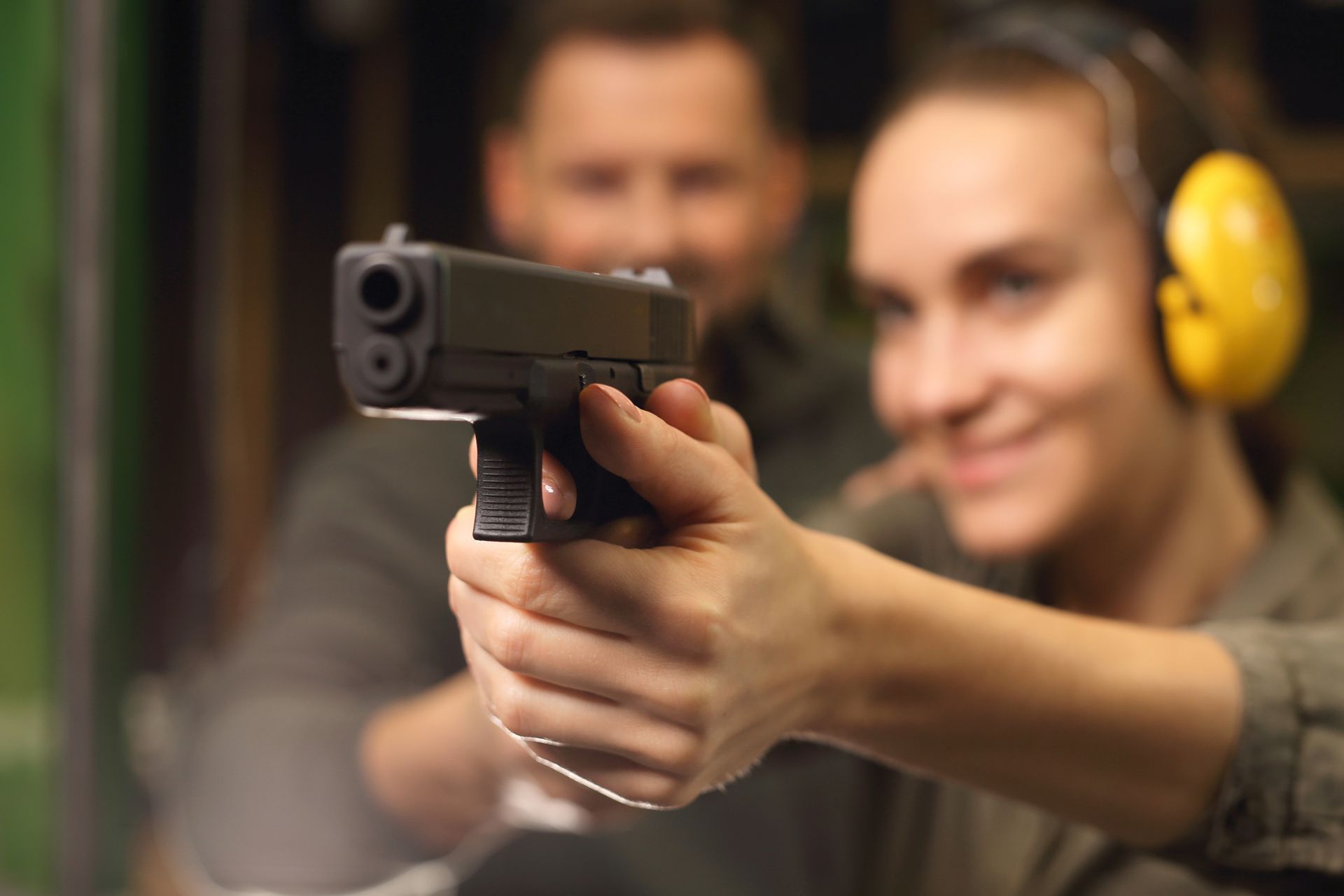 Woman aiming a handgun at a shooting range, with a man in the background, both wearing ear protection.