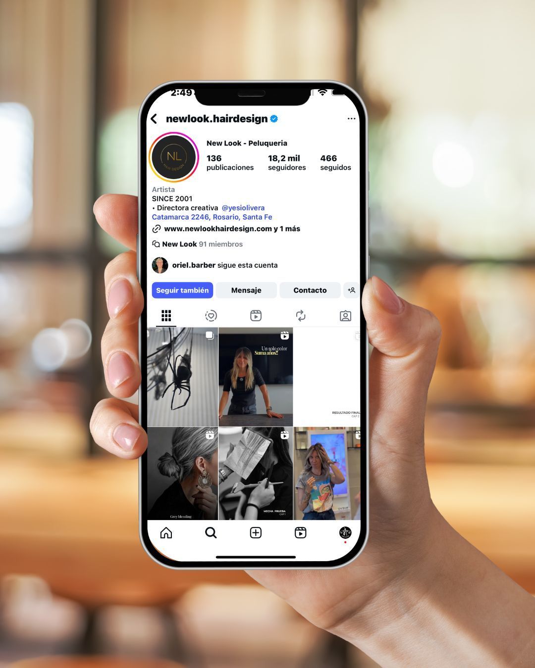 Unas manos sostienen un teléfono, tomando una foto de una decoración iluminada, junto a un ordenador portátil y un vaso de agua sobre una mesa blanca.