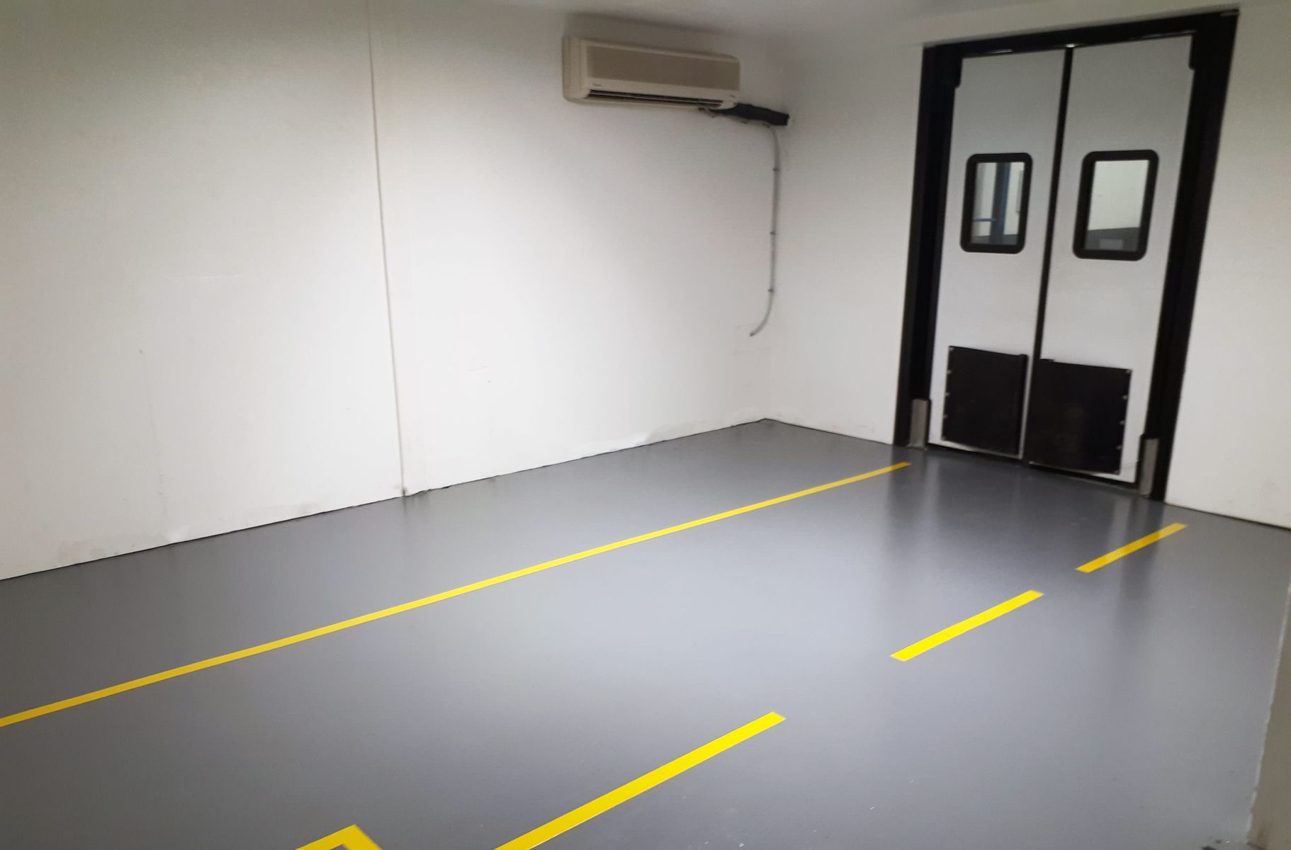 Pièce vide au sol gris, avec portes doubles, marquages ​​jaunes au sol, murs blancs et climatiseur.