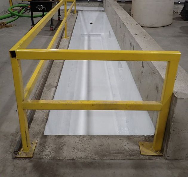 Dans un contexte industriel, une rambarde jaune entoure une tranchée en béton à revêtement blanc.