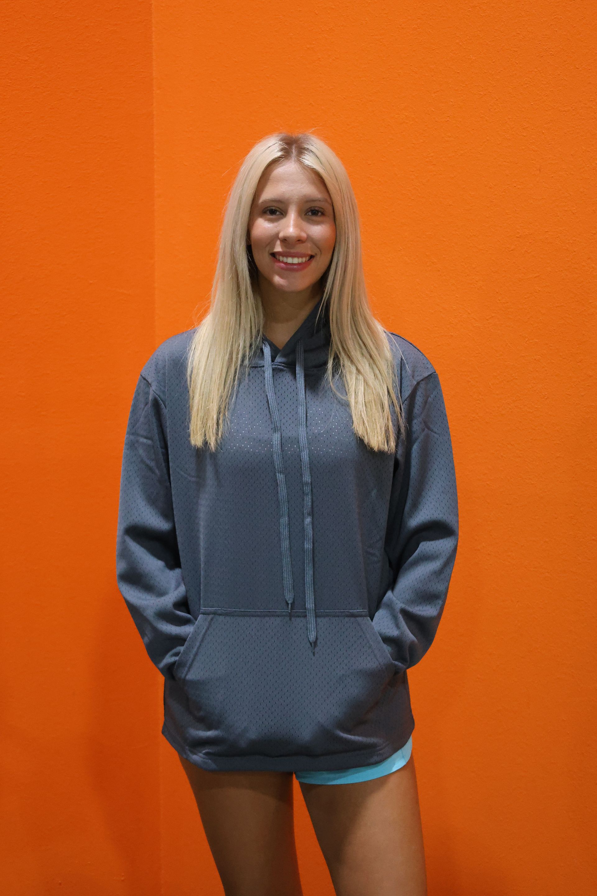 A woman wearing a grey hoodie and shorts is standing in front of an orange wall.
