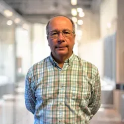 A man in a plaid shirt and glasses is standing in a hallway.