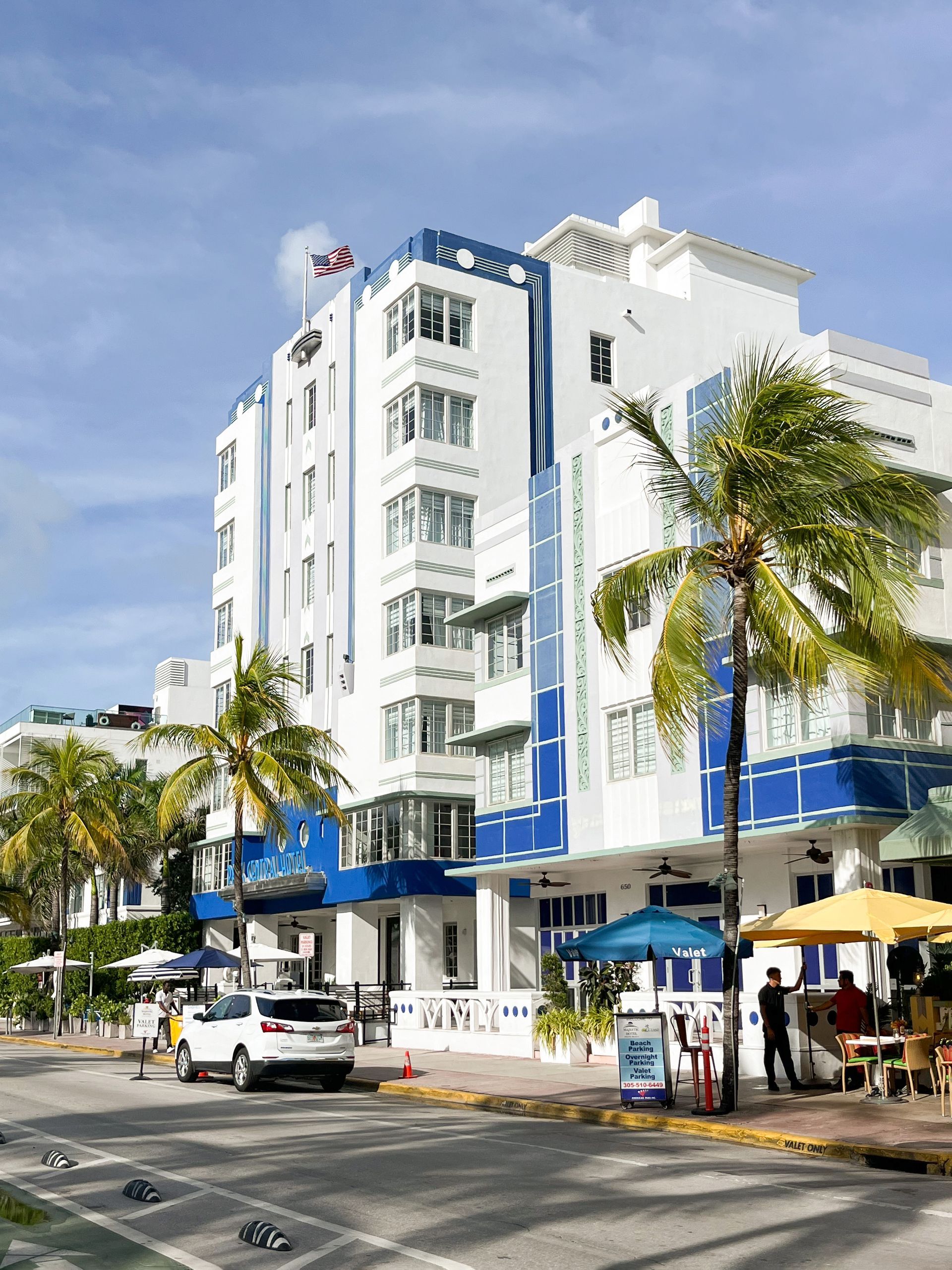 A bright blue and white Art Deco hotel in Miami, with palm trees, a car, and people on the sidewalk.