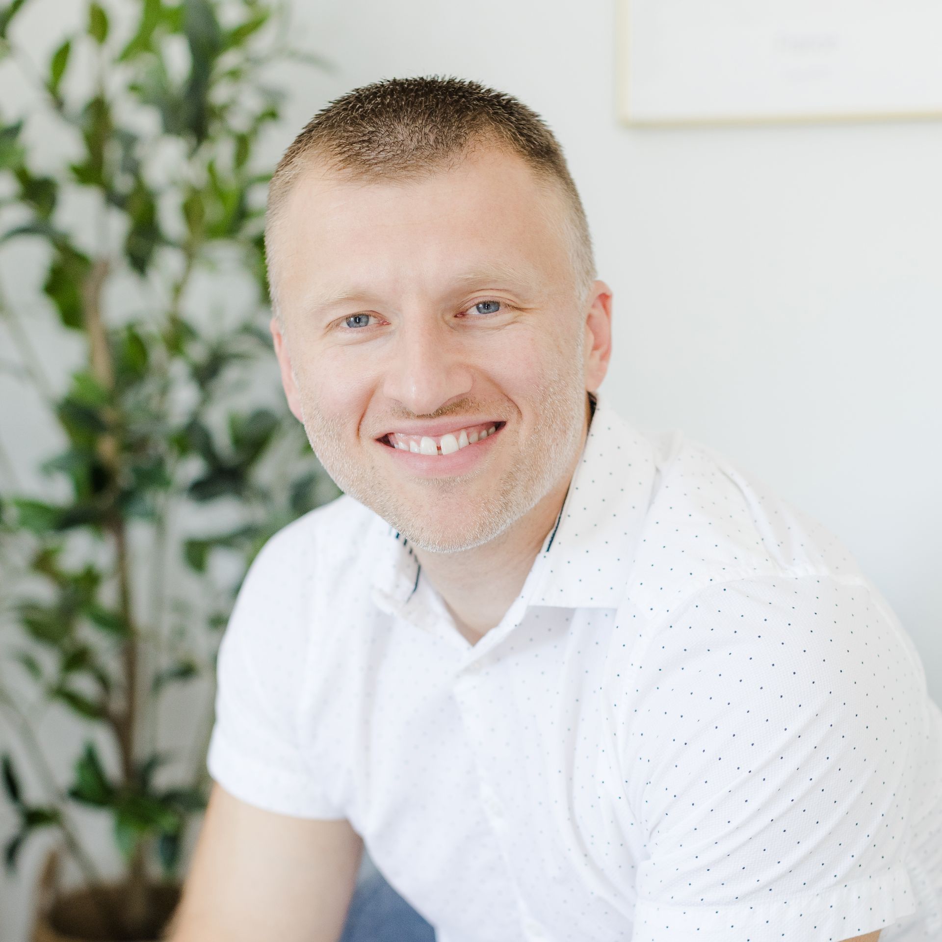 Man with short hair, smiling in a white shirt, indoor setting, green plant in the background.