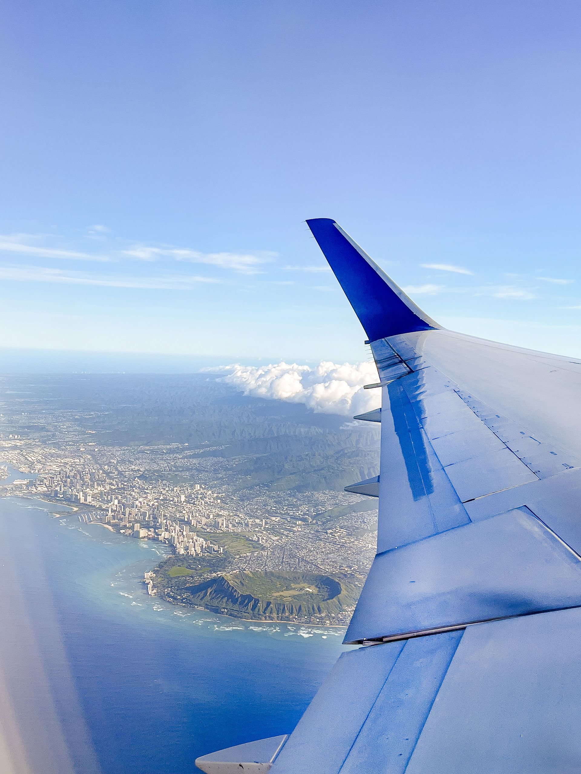 View from airplane window over a coastal city, wing in foreground; blue water and sky, clouds.