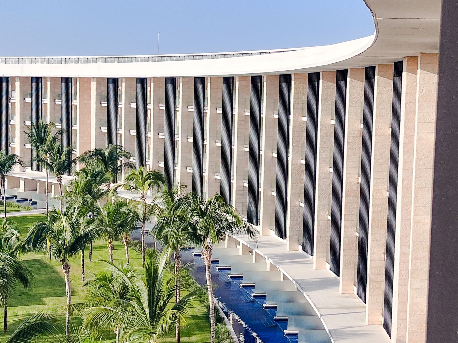 Exterior view of a modern building with vertical columns, palm trees, and a blue water feature.