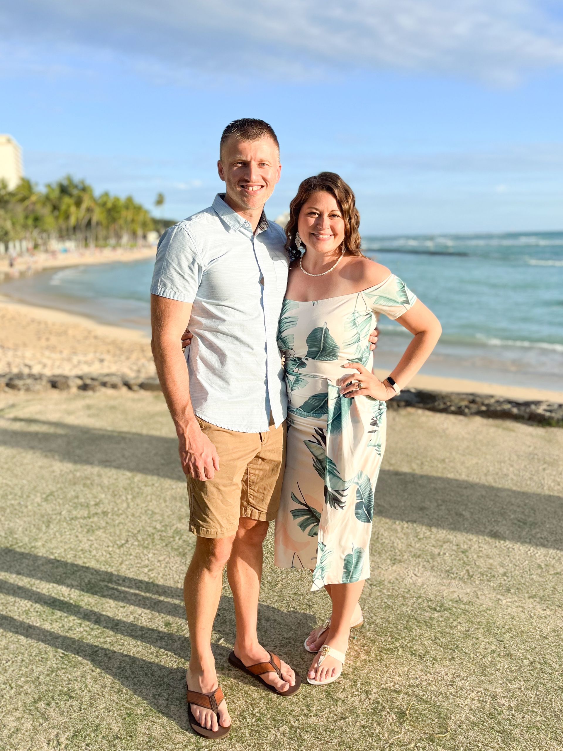 Couple on a beach, man in shorts and shirt, woman in dress. Beach and ocean in the background, smiling.