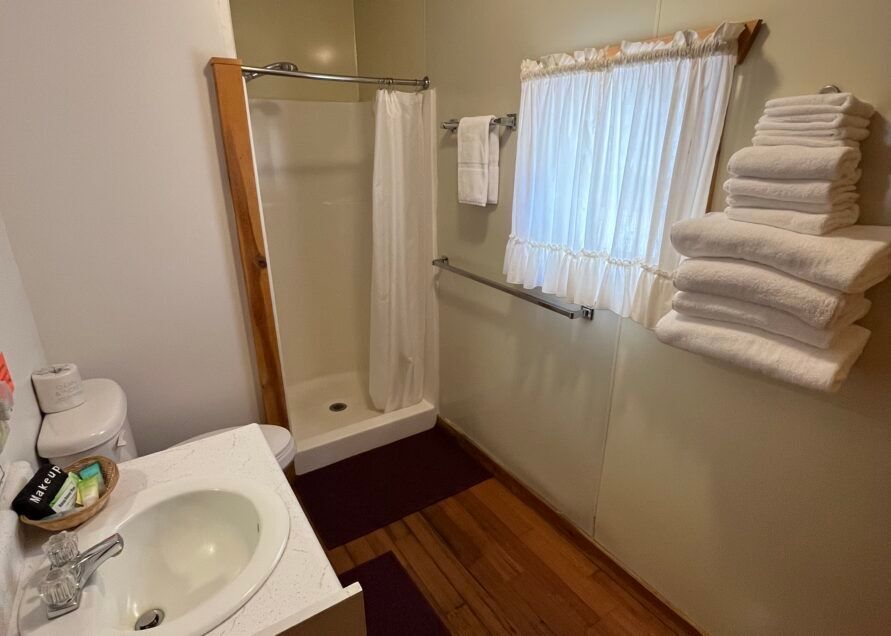 Small bathroom with a shower, sink, toilet, and window. White towels are stacked on the wall. Wooden floor and beige walls.