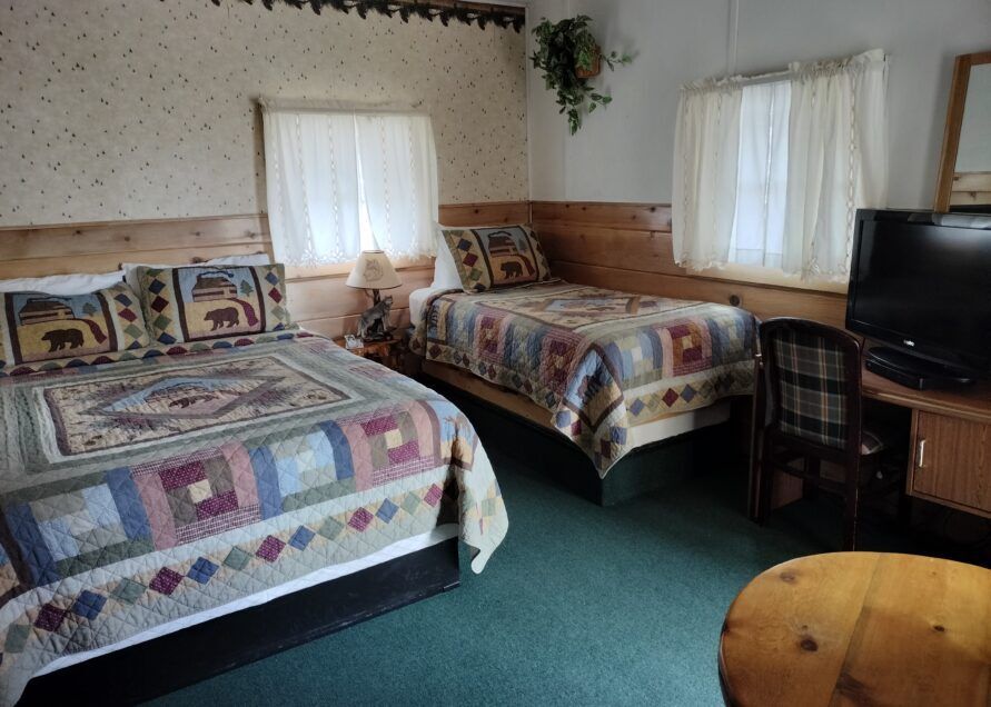 A rustic motel room with a double bed and a single bed, patterned quilts, two windows, and a TV on a desk.