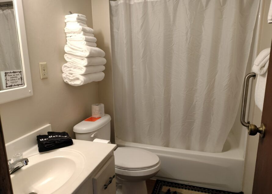 Bathroom with a white tub/shower, toilet, sink, and stacked white towels.