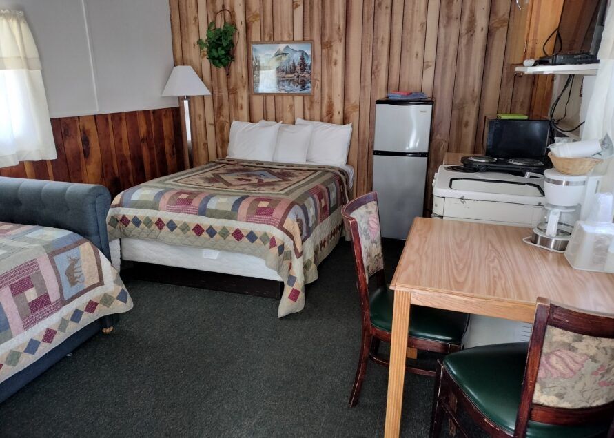 A rustic motel room with a double bed, single bed, small fridge, desk, and wood-paneled walls.