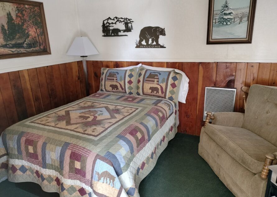 A rustic bedroom with a bed, armchair, artwork, and bear-themed decor. Walls are wood-paneled with green carpet.