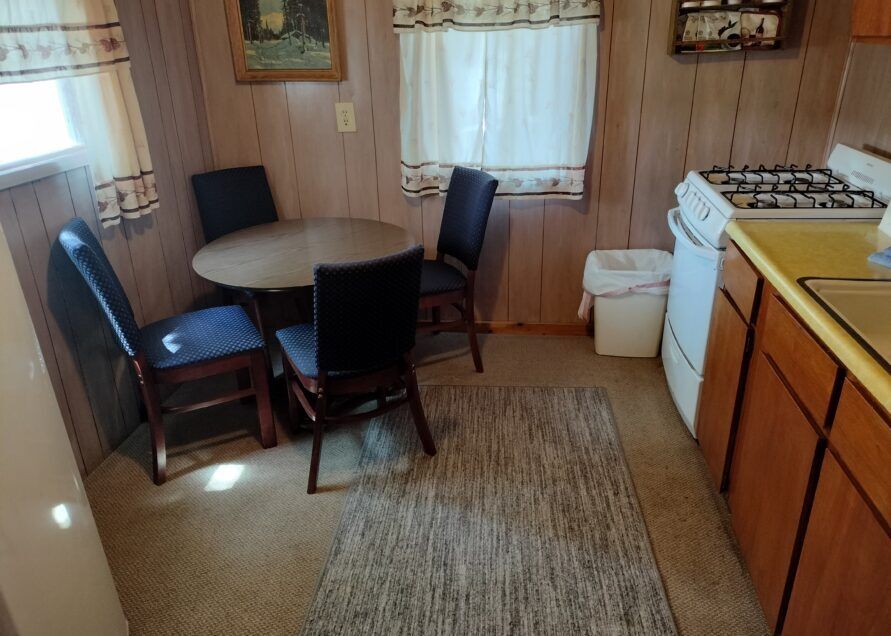 A small, wood-paneled kitchen with a round table and four chairs. A rug sits in front of the stove.