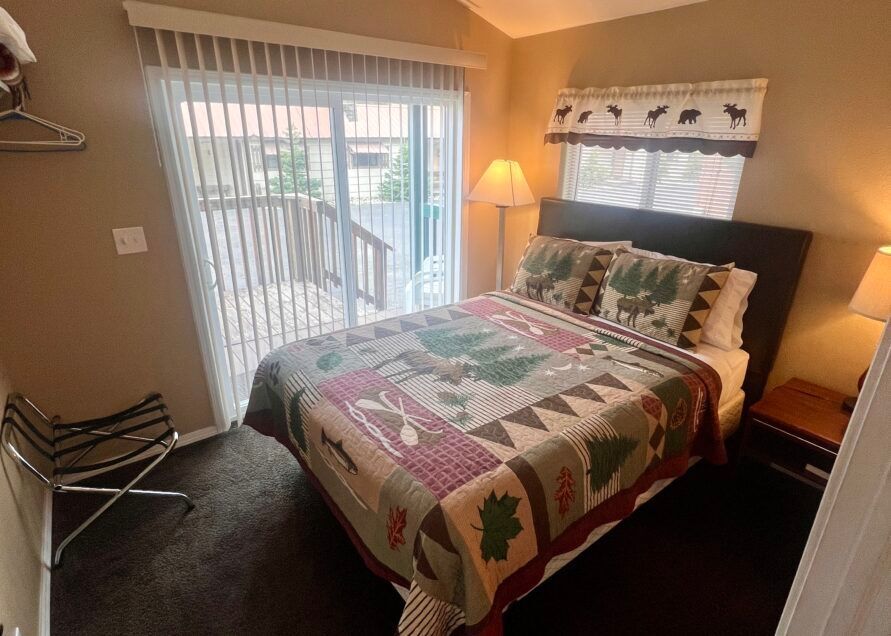 Bedroom with a bed covered in a quilt, sliding glass door to a deck, small chair, and nightstand.
