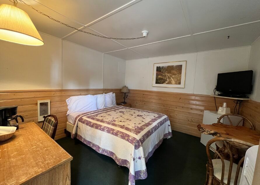 A small motel room featuring a bed with a purple floral quilt, a TV, and a wooden desk.