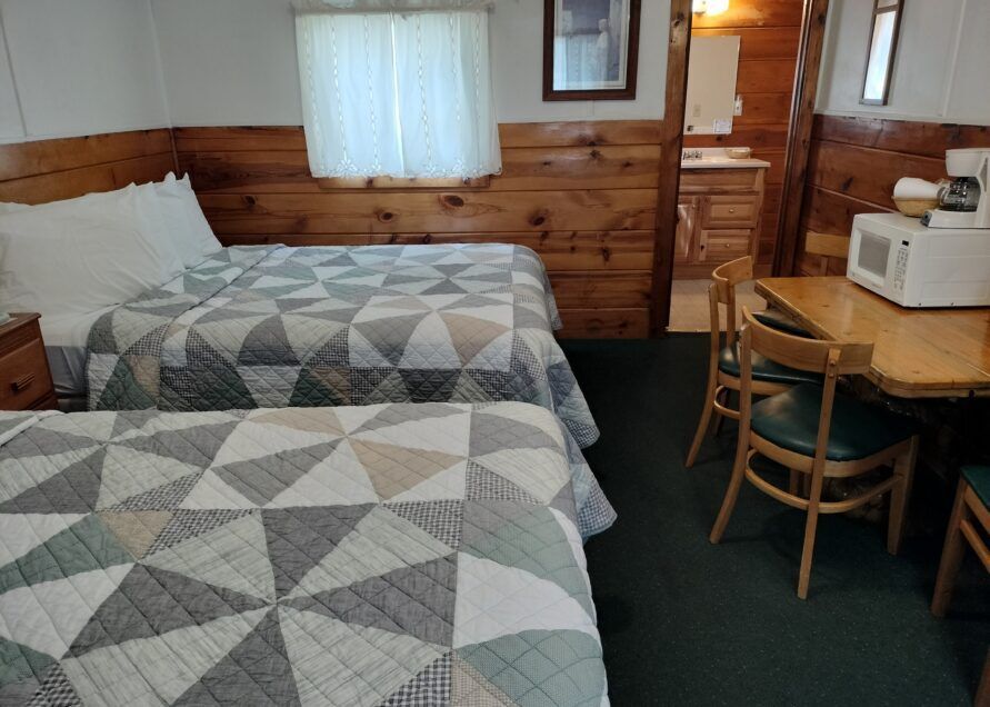 A motel room with two beds, a microwave, small table with chairs, and a bathroom. The walls are wood-paneled.
