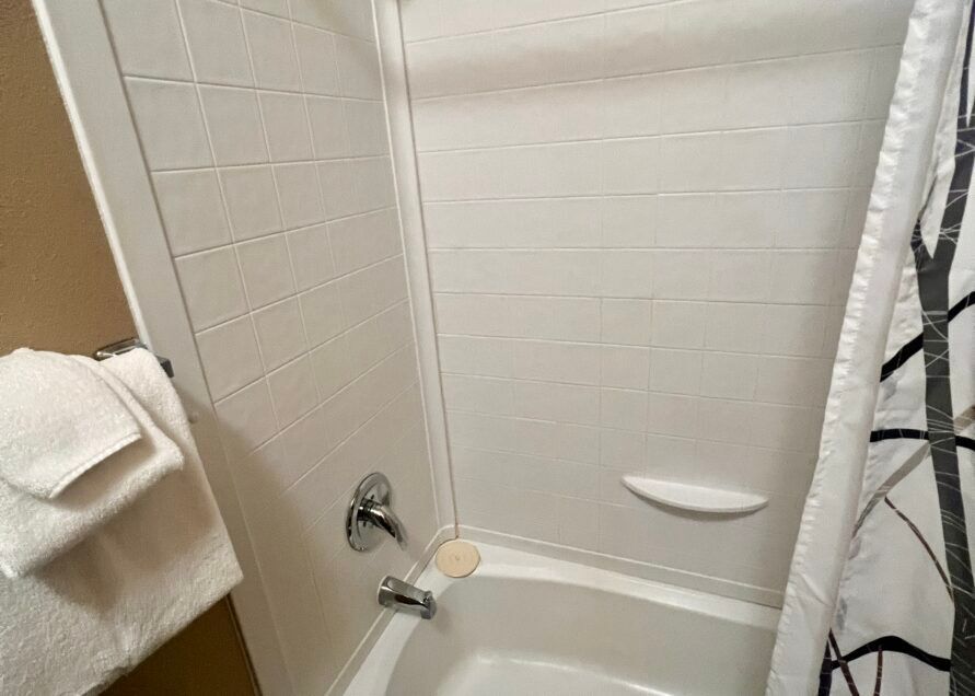 White-tiled shower with a bathtub, soap dish, and shower curtain. Two white towels hang on a rack to the left.