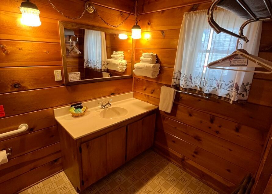 Cozy bathroom with wooden walls, sink, mirror, towels, and window with curtain. Two lights illuminate the space.