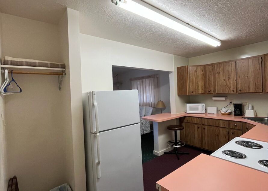 A small motel kitchenette with white fridge, pink countertops, and wooden cabinets. A doorway leads to a bedroom with a bed.