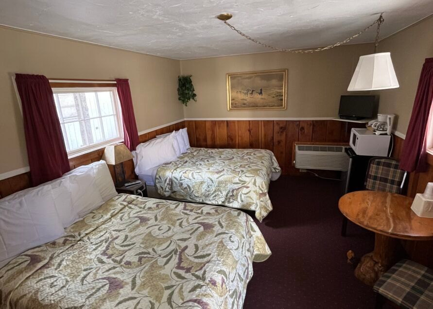 A motel room with two double beds, a small table, a window, and a TV. Beige walls, dark red curtains, and floral bedspreads.