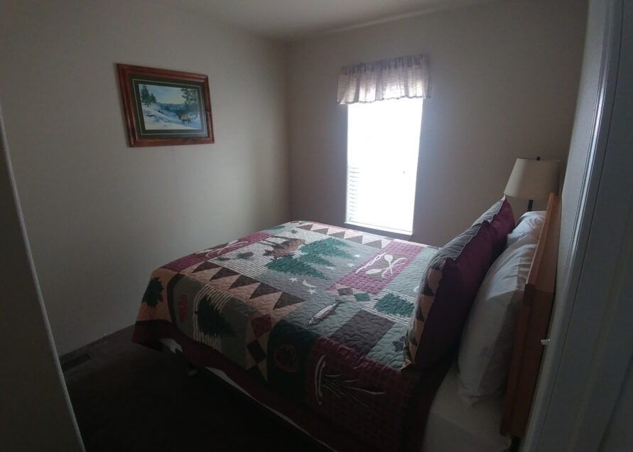 Bedroom with a bed covered with a quilt, a landscape painting on the wall, and a window with a curtain.