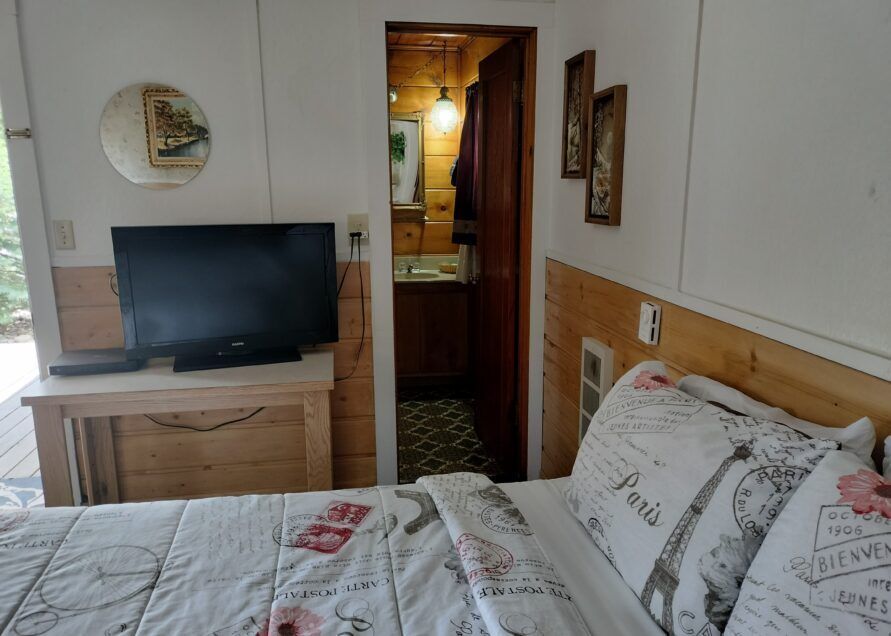 Cozy bedroom with a bed, TV, and a view into a small bathroom. Wooden paneling and decorations add to the cabin-like feel.