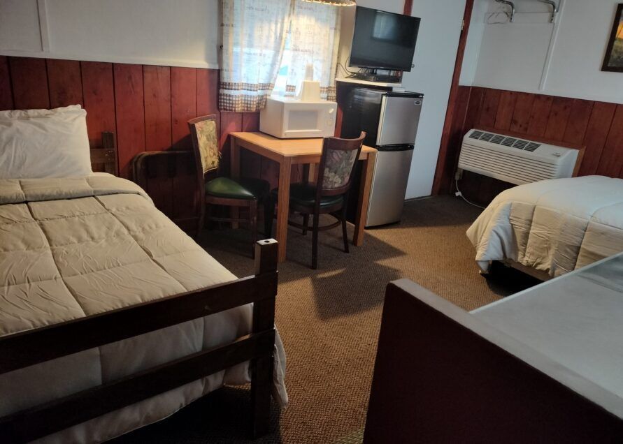 A small motel room with two beds, a table with chairs, a microwave, and a mini-fridge. Walls are dark red.