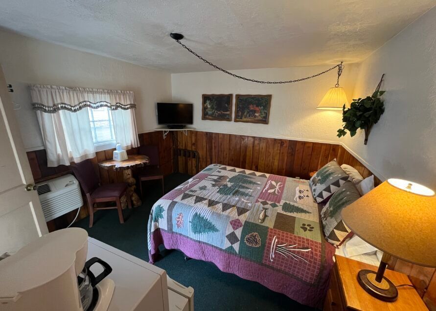 Small motel room with a bed, TV, and small table. The room features a quilt, wooden paneling, and a warm lamp.