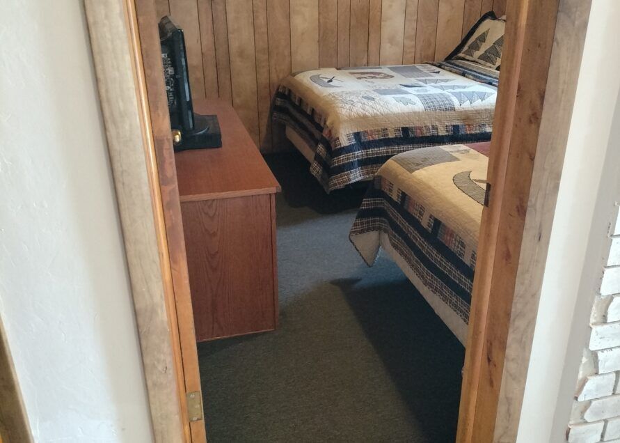 A small bedroom with two beds, a dresser, and a TV seen through a doorway. The walls are wood-paneled.