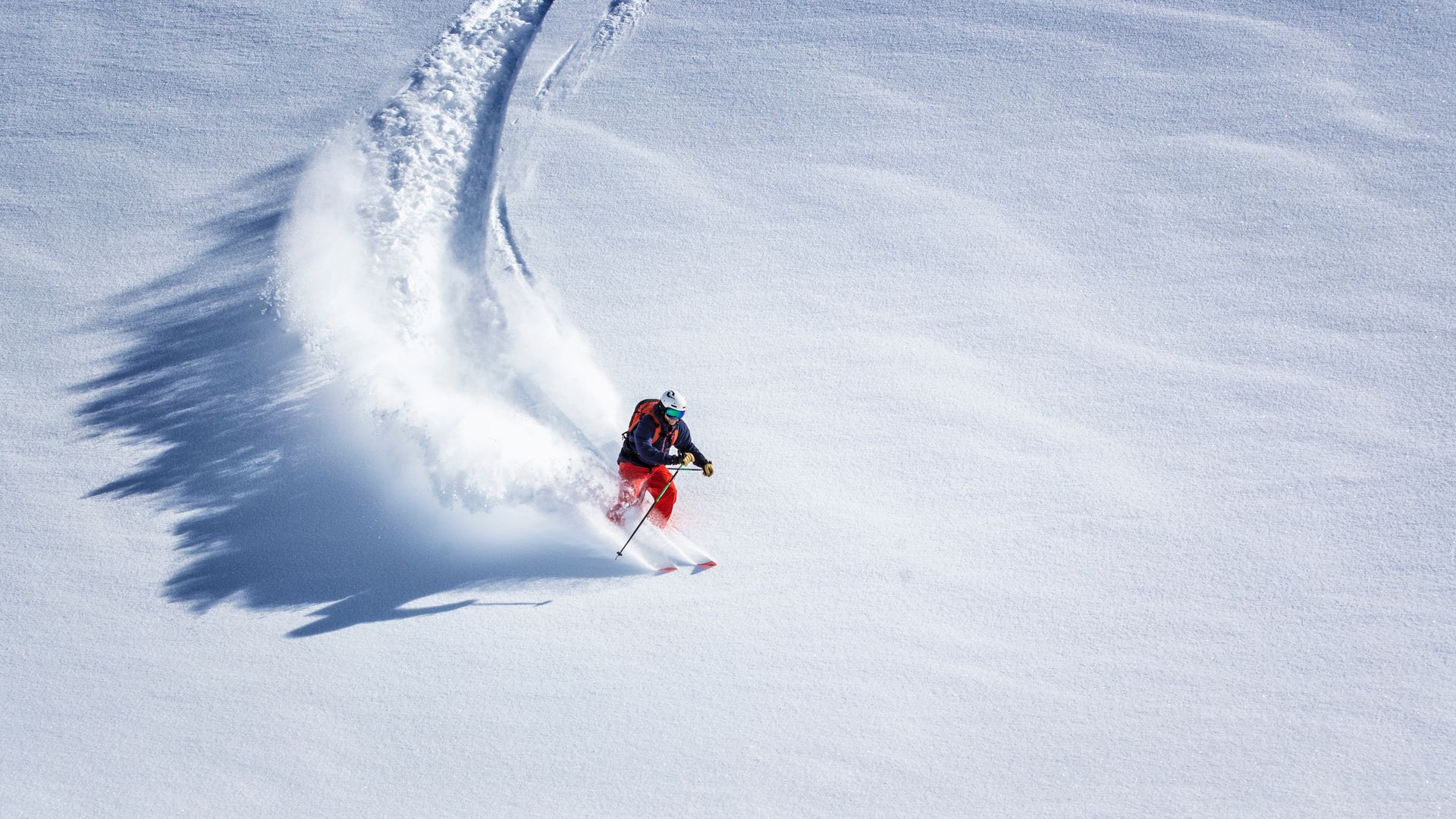 Skier carving through deep powder snow, creating a large white cloud. Set in a snowy, mountainous terrain.