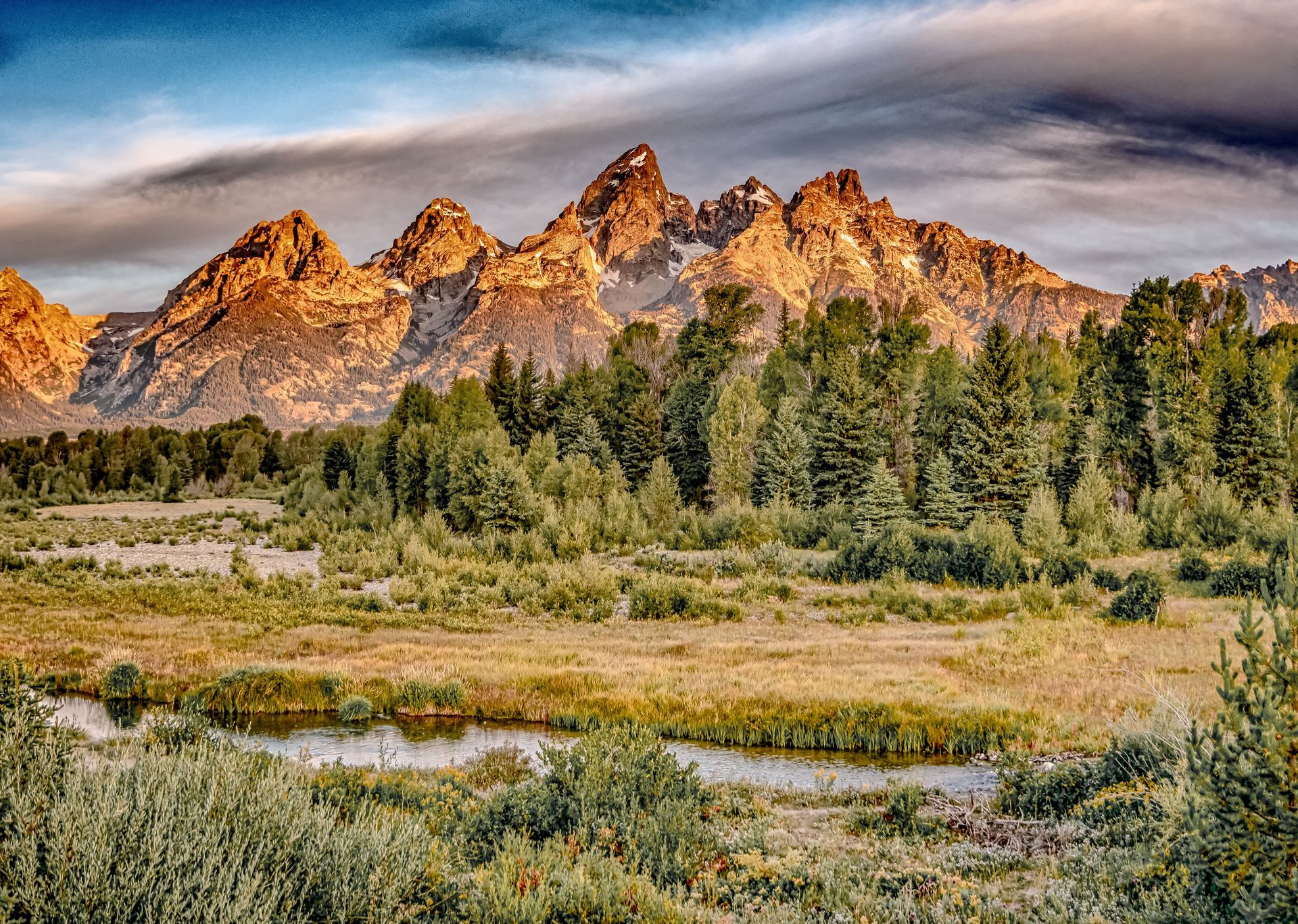 Mountains at sunset, bathed in warm light, rise above a forest and marshy landscape.