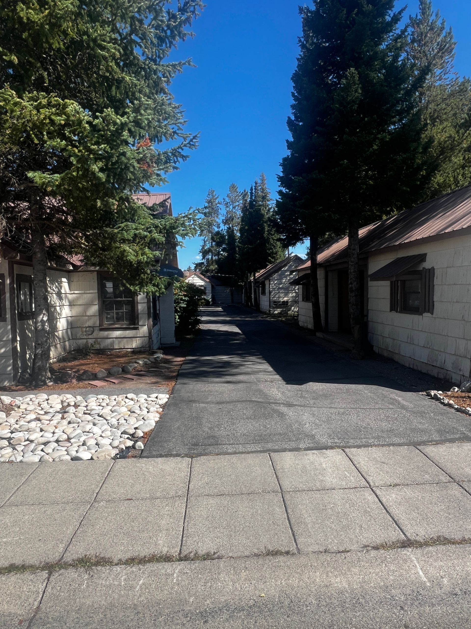 Narrow street lined with small buildings, trees, and blue sky. Concrete sidewalk leads to a dark asphalt path.