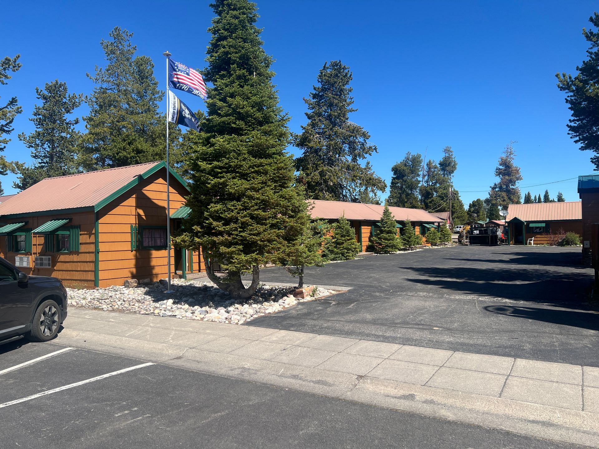 Lodge-style cabins with green roofs and an American flag in a sunny parking lot under blue sky