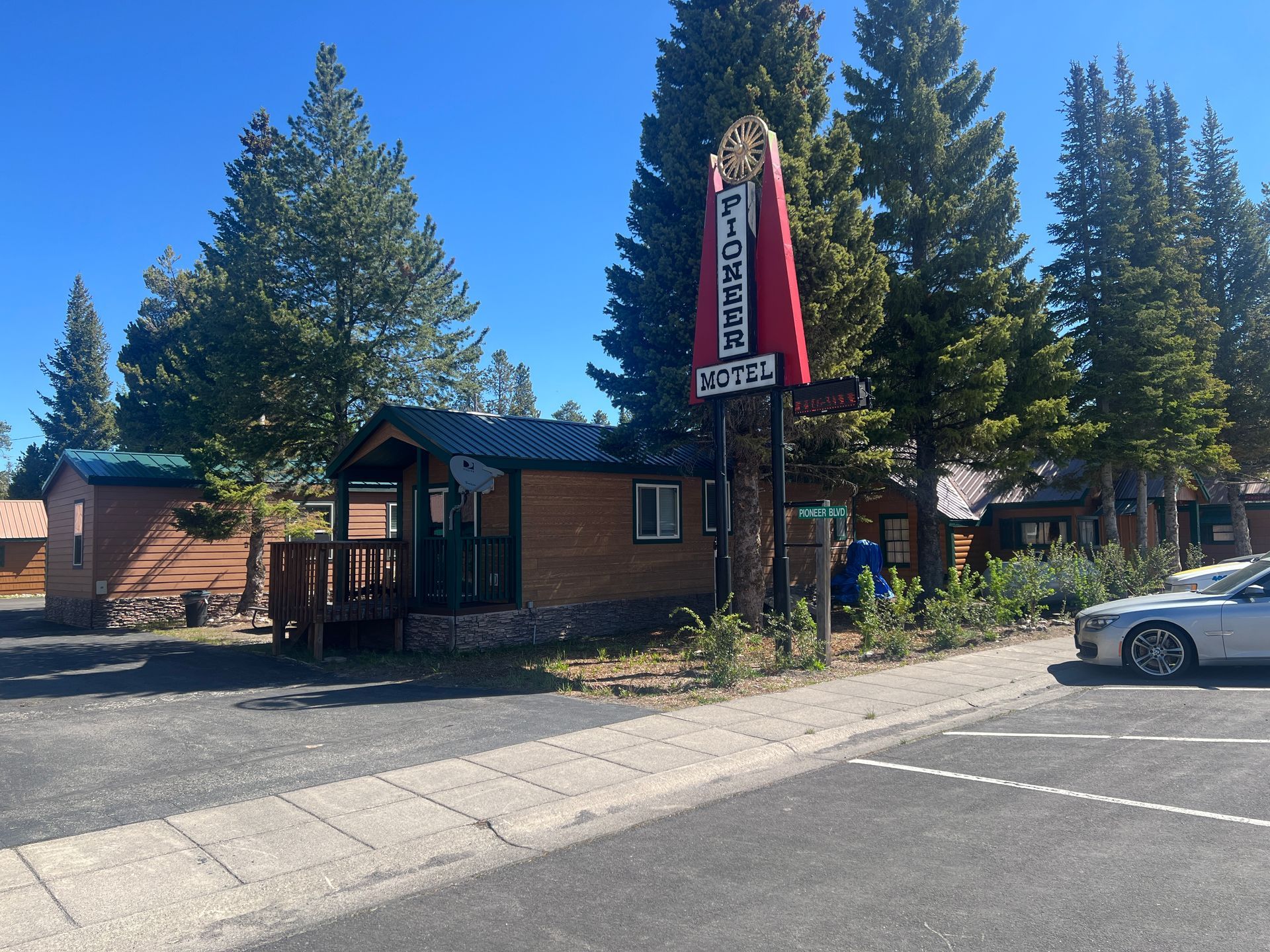 Small roadside lodge with red vertical sign, pine trees, and parked cars on a sunny street.