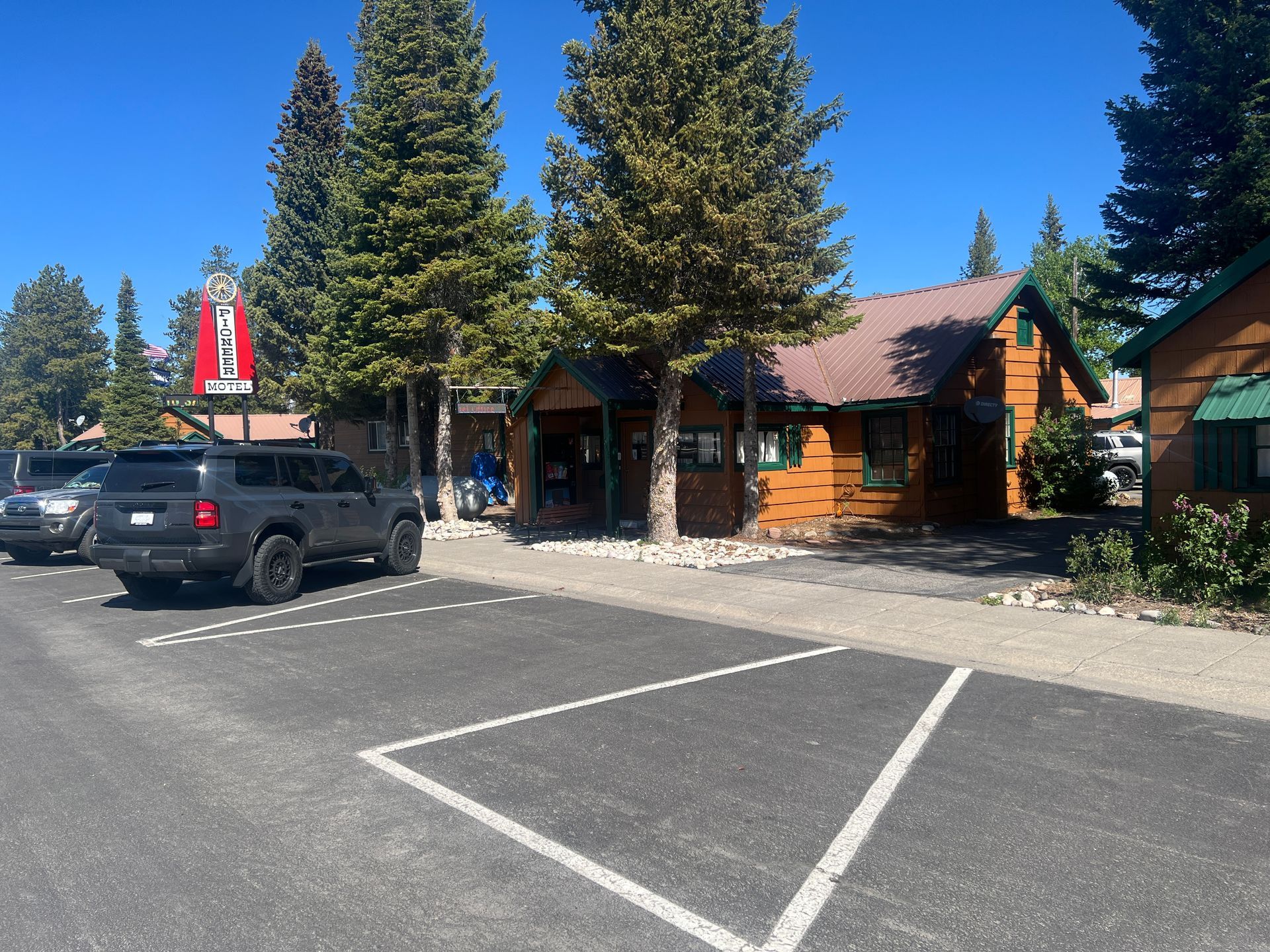 Parking lot beside rustic cabins with a black SUV and tall trees under a clear blue sky