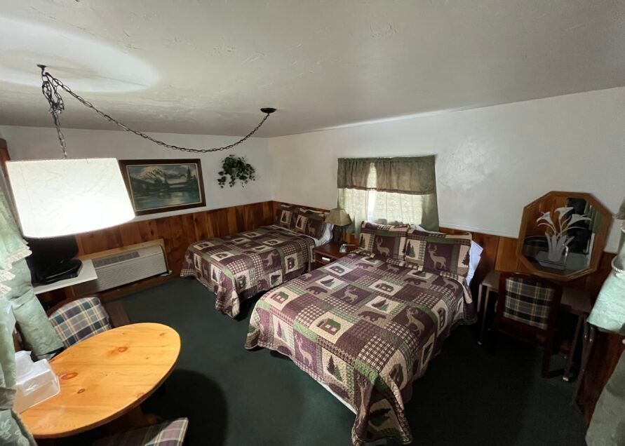 A rustic motel room with two double beds covered in a bear-themed quilt, a small table, and a vintage mirror.