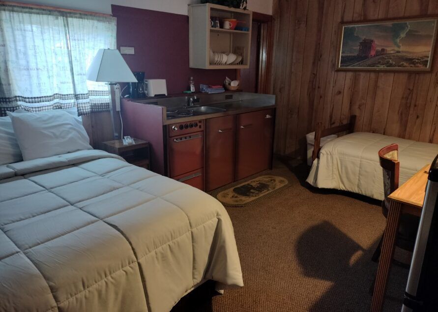 A small motel room with a double bed, single bed, kitchenette, and artwork on the wood-paneled walls.