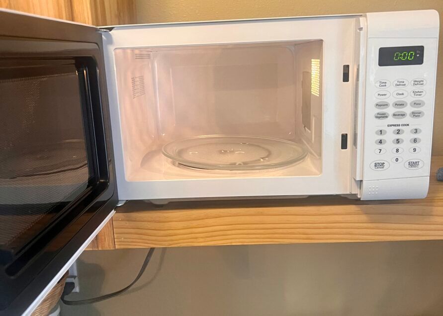 An open, empty white microwave sitting on a wooden shelf. The glass turntable is visible inside.