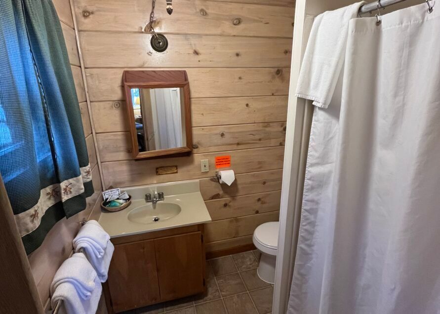 A small bathroom with a wooden wall. It has a sink, mirror, toilet, and shower with a white curtain.