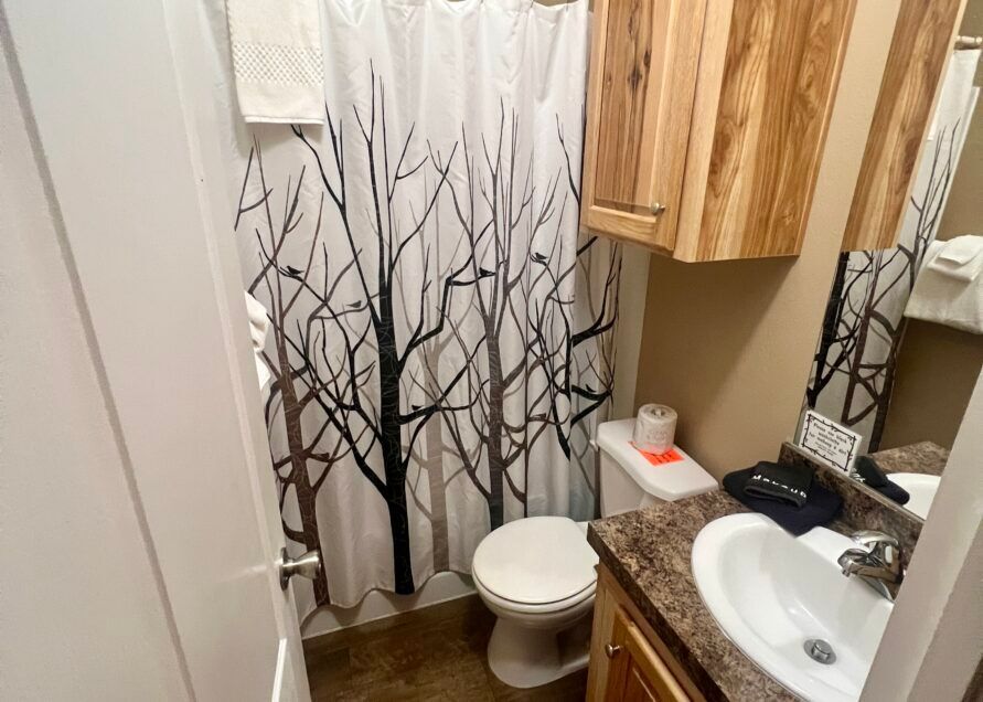 Small bathroom with a tree branch-patterned shower curtain, toilet, sink, and wooden cabinets.
