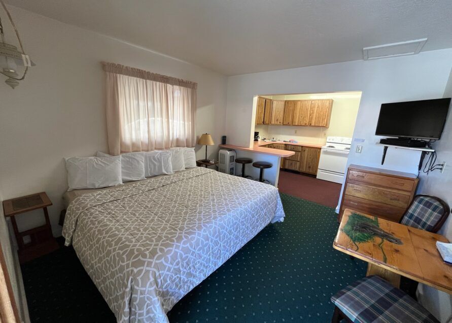 Cozy motel room with a queen bed, small kitchen, and a TV. Features a wooden desk and green carpet.