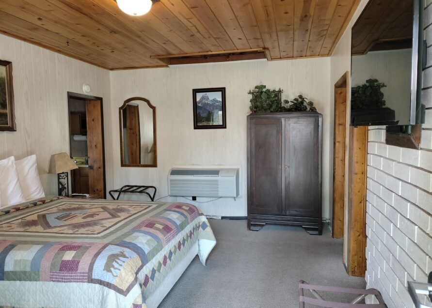 A motel room with a bed, wardrobe, mirror, and air conditioner, featuring a wood-paneled ceiling and quilt.