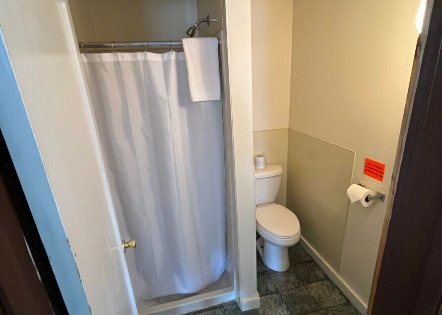 A small bathroom with a shower, toilet, and toilet paper roll. The walls are beige, and the floor is tiled.