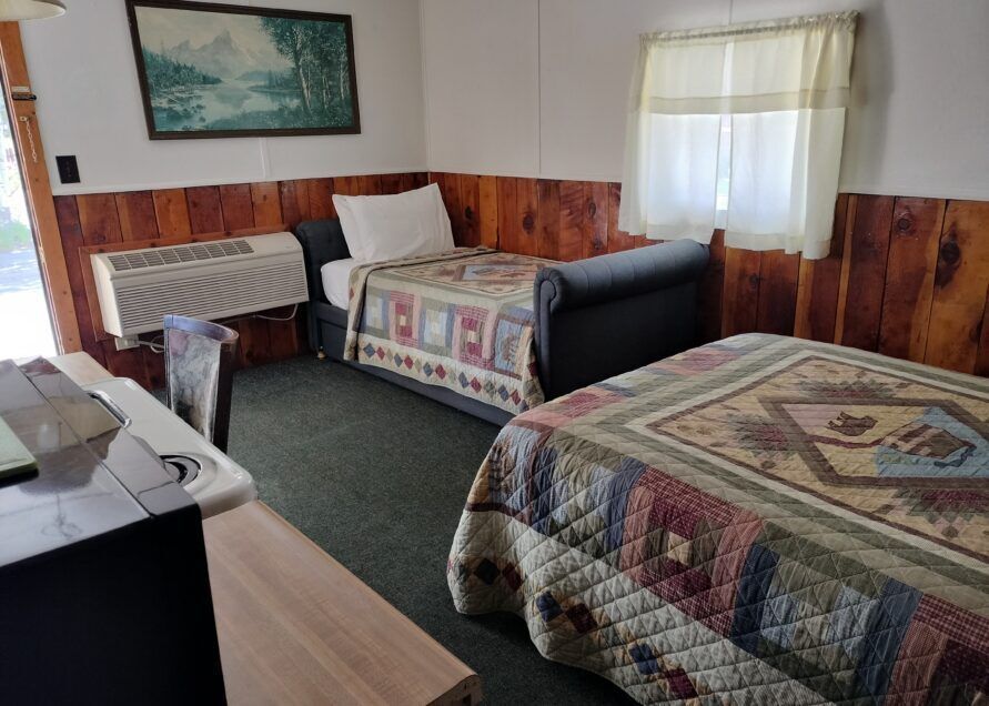 Small motel room with two beds, wood paneling, and a window. A painting hangs above one bed.