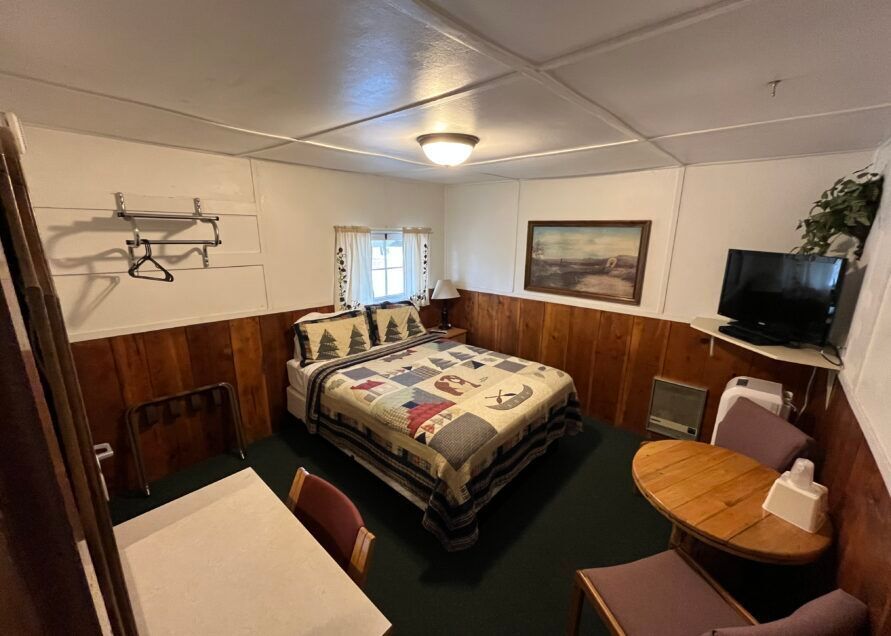 A small, cozy motel room.  It features a bed with a quilt, a table with chairs, a TV, and wood paneling.