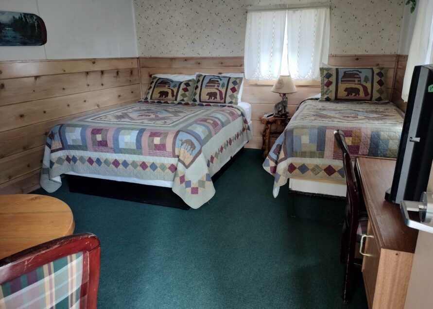 A motel room with two beds covered in quilts, a desk with a computer, and a round table. The room has wood paneling.