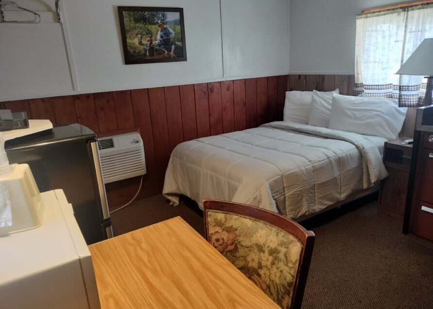 A small motel room featuring a bed with white bedding, a small desk, a mini-fridge, and dark wood paneling.